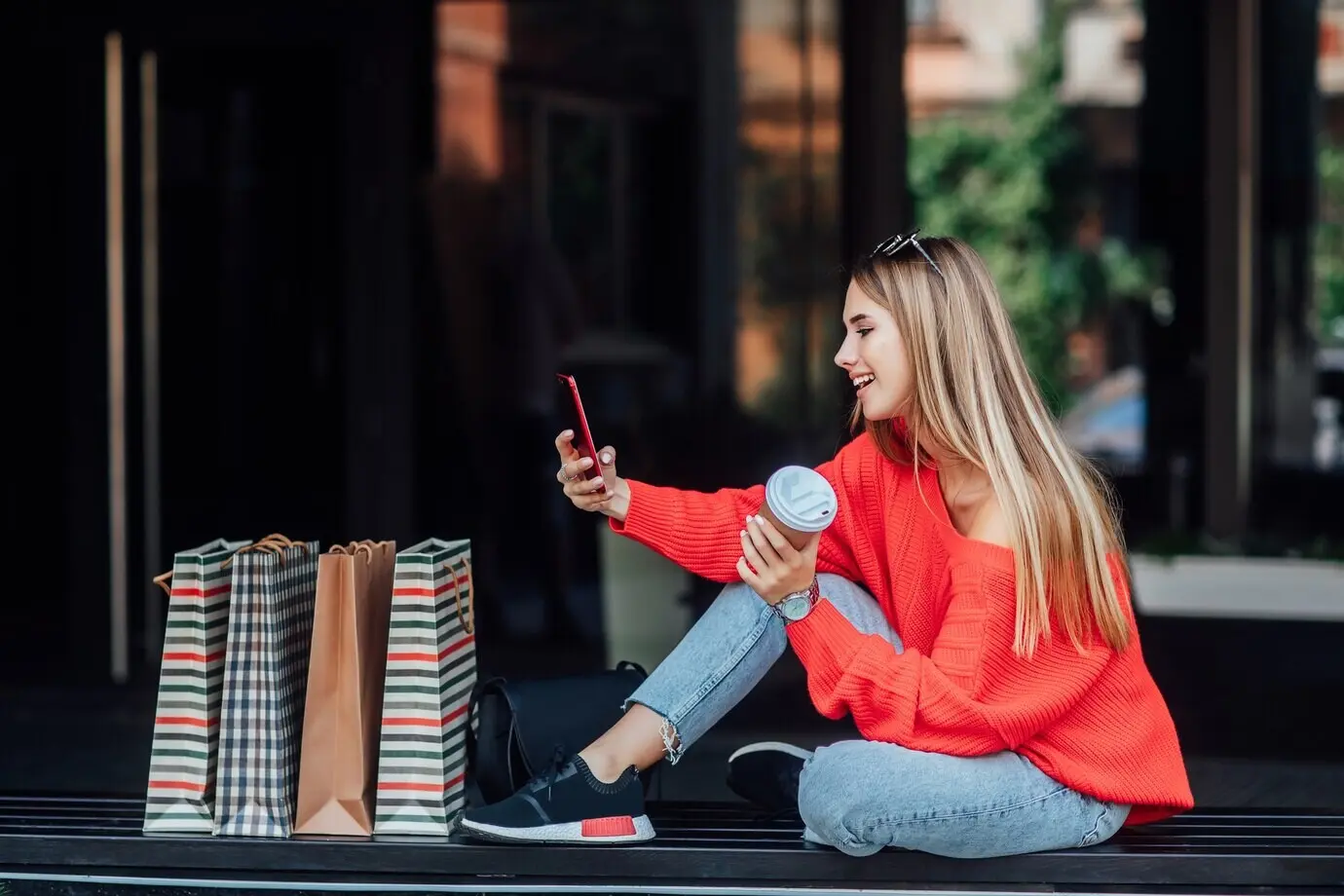 A beautiful blonde woman sits on the street, surrounded by shopping bags, looking at her phone.